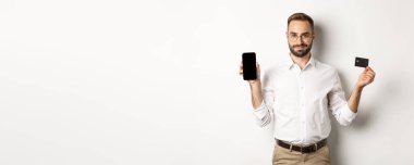 Cep telefonu ve kredi kartı gösteren yakışıklı sakallı bir adam. İnternetten alışveriş yapıyor, beyaz arka planda duruyor..