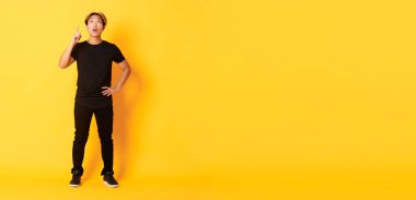 Uzun boylu, düşünceli, ilgi çekici sarışın Asyalı adam parmağını kaldırıyor, yukarı bakıyor ve eğleniyor görünüyor, sarı arka plan..