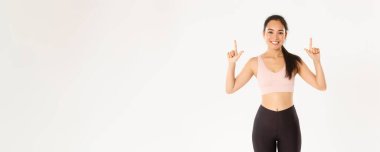Spor, refah ve aktif yaşam tarzı konsepti. Gülümseyen ince ve tatlı Asyalı fitness kızı, parmaklarını gösteren bayan sporcu, egzersiz malzemeleri satış promosyonu veya spor salonu üyeliği teklifi.