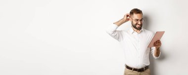 Kafası karışmış erkek yönetici dijital tablete şaşkın bakıyor, kafasını kaşıyor, beyaz arka planda duruyor..