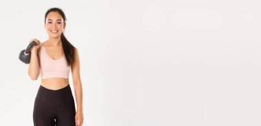 Spor, refah ve aktif yaşam tarzı konsepti. Güçlü ve ince Asyalı fitness kızı, kettlebell tutan bayan sporcu ve kaygısız görünen, spor salonunda kaslı, beyaz arka planda duran.