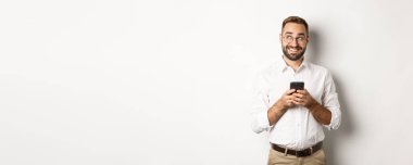 Düşünceli yakışıklı yönetici cep telefonu kullanıyor ve mesajlara cevap vermeyi düşünüyor, sol üst köşeye bakıyor ve gülümsüyor, beyaz arka planın üzerinde duruyor..