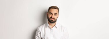 Şüpheci sakallı, hoşnutsuz, bir şeyden hoşlanmayan, beyaz arka plana karşı gönülsüz duran adamın yakın çekimi..