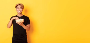 Mutlu, yakışıklı Koreli adam. Mutlu bir şekilde gülümsüyor. Film ya da TV dizisi izliyor, patlamış mısır yiyor, sarı arka planda duruyor..