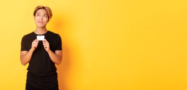 Düşünceli gülümseyen Asyalı adam kredi kartı gösterirken düşünüyor, sol üst köşeye bakıyor rüya gibi, sarı arka plan.