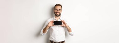 Gözlüklü yakışıklı sakallı adam, cep telefonu uygulamasını gösteriyor, akıllı telefon ekranını gösteriyor, beyaz arka planda duruyor..