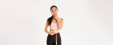 Fitness, sağlıklı yaşam tarzı ve refah konsepti. Düşünceli Asyalı kadın spor hocasının portresi, daha fazla kilo vermeyi düşünen sporcu kadın, spor yaptıktan sonra bel ölçüsü, beyaz arka plan..