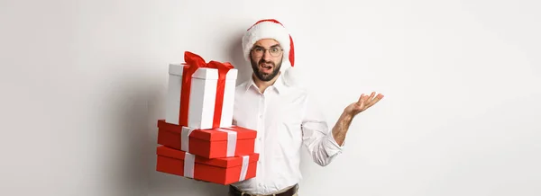 メリークリスマス休暇のコンセプト 白い背景にサンタの帽子に立って困惑しながら クリスマスの贈り物を保持しながら 男は混乱して見える — ストック写真