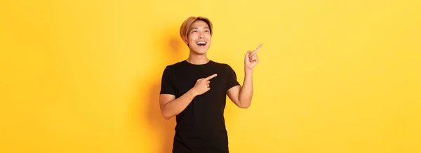 幸せなハンサムなアジアの男の肖像画で黒Tシャツ 指差し指と満足笑顔で左上隅を見て 黄色の背景 — ストック写真