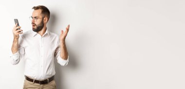 Konuşmadan sonra cep telefonuna şaşkın şaşkın bakan bir adam, beyaz arka plan üzerinde şaşkın şaşkın duruyor. Boşluğu kopyala