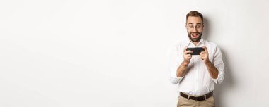 Adam heyecanlı ve şaşırmış görünüyor, akıllı telefonu yatay olarak tutuyor, beyaz arka planda duruyor..