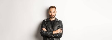 Çekici maço deri ceketli, gülümseyen, kendine güvenen, göğsünde çapraz ellerle beyaz arka planda bir adam..
