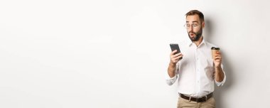 Kahve içen ve cep telefonundaki mesaja şaşıran bir iş adamı beyaz arka planda şaşkın şaşkın dikiliyor..