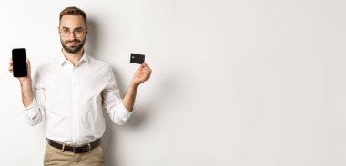 Cep telefonu ve kredi kartı gösteren yakışıklı sakallı bir adam. İnternetten alışveriş yapıyor, beyaz arka planda duruyor..