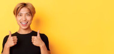 Tatmin olmuş, siyah tişörtlü mutlu Asyalı adam. Onayını veriyor, gülümsüyor ve katılıyor. Sarı arka planda duruyor..