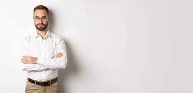 Gözlüklü kendine güvenen işadamı kameraya bakıyor, göğsünde çapraz kollar, beyaz arka planda duruyor..