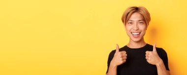 Tatmin olmuş, siyah tişörtlü mutlu Asyalı adam. Onayını veriyor, gülümsüyor ve katılıyor. Sarı arka planda duruyor..