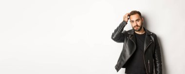 Siyah motorcu ceketi giyen yakışıklı maço adam, saç kesimine dokunuyor ve havalı görünüyor, beyaz arka plan..