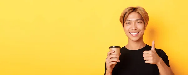 満足したアジアのハンサムな男のクローズアップは カフェをお勧めします コーヒーのカップを保持し 承認で親指アップを示す 黄色の背景に満足笑顔 — ストック写真