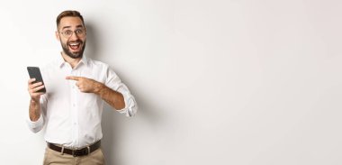 Adam heyecanlı görünüyor ve cep telefonunu işaret ediyor, iyi bir online teklif sunuyor, beyaz arka planda duruyor..