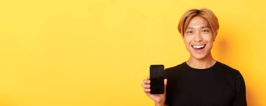 Akıllı telefon ekranı gösteren ve gülümseyen yakışıklı Koreli adamın yakın plan fotoğrafı. Sarı arka planda duran mobil uygulamayı tavsiye et..