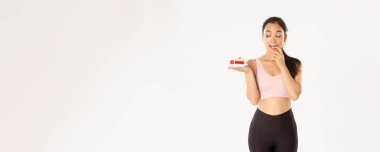 Aktif yaşam tarzı, fitness ve refah konsepti. Tatlı esmer Asyalı sporcu kız, lezzetli pastalar yemek istiyor ama diyette olmak, vücut ağırlığı ve kalorilerle ilgilenmek, beyaz arka plan..