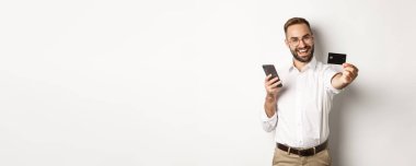 İş ve çevrimiçi ödeme. Akıllı telefon tutarken kredi kartını gösteren heyecanlı bir adam beyaz arka planda tatmin olmuş bir şekilde duruyor..