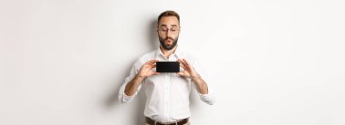 Mobil ekran gösteren neşeli yakışıklı adam, çevrimiçi web sitesine bakıyor, beyaz arka planda duruyor..
