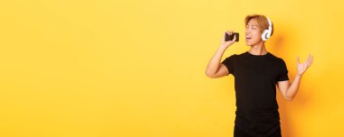 Kulaklıklı neşeli çekici Koreli adam, karaoke uygulaması oynuyor, cep telefonu mikrofonuna şarkı söylüyor, sarı arka planda duruyor..