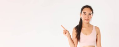 Spor, refah ve aktif yaşam tarzı konsepti. Kurnaz, düşünceli, gülümseyen Asyalı sporcu, seçimini yapan fitness kızı, meraklı bir gülümsemeyle mutlu görünüyor, sol üst köşeyi işaret ediyor..