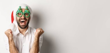 Noel 'i ya da yeni yılı kutlayan, Noel Baba gözlüğü takan, Noel Baba şapkası takan, sevinen ve sevinen, beyaz arka planda duran bir adamın yakın çekimi..