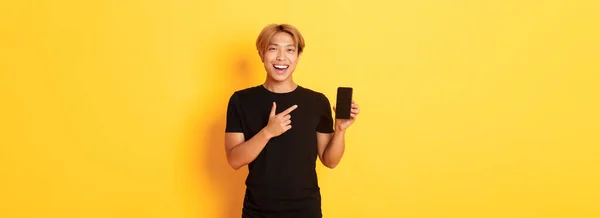 満足した笑顔アジアの男の肖像画ブロンドの髪 スマートフォンの画面で面白がって指差す指を見て 立って黄色の背景 — ストック写真