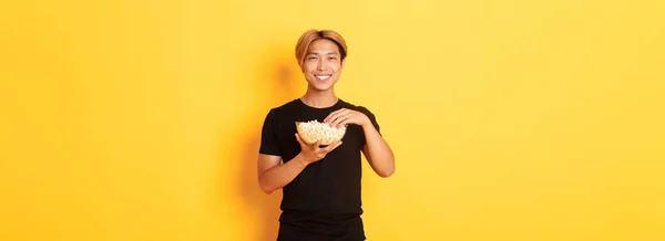 楽しい映画やテレビシリーズを見て ポップコーンを食べて 立って黄色の背景として幸せな笑顔ハンサムな韓国の男を喜ばせます — ストック写真