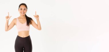 Spor, refah ve aktif yaşam tarzı konsepti. Gülümseyen ince ve tatlı Asyalı fitness kızı, parmaklarını gösteren bayan sporcu, egzersiz malzemeleri satış promosyonu veya spor salonu üyeliği teklifi.