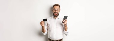 İş ve çevrimiçi ödeme. Heyecanlı adam cep telefonu ve kredi kartıyla ödeme yapıyor, şaşkın bir şekilde gülümsüyor, beyaz arka planda duruyor..