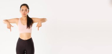 Spor, refah ve aktif yaşam tarzı konsepti. Surat asan ve hayal kırıklığına uğramış Asyalı antrenman kızı, aktif giysili bayan sporcu üzgün, kötü bir şeye işaret eden ve ona tepeden bakan, beyaz arka plan.