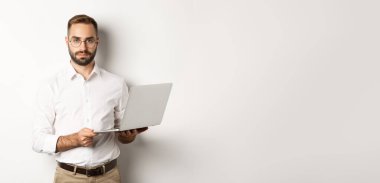 İş için. Bilgisayarda çalışan yakışıklı bir yönetici, elinde bilgisayarla kameraya bakıyor, beyaz arka planda duruyor..