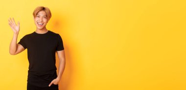 Siyah kıyafetli yakışıklı Asyalı adamın portresi, merhaba demek ve gülümsemek için el sallıyor, birini selamlıyor, sarı arka planda duruyor..