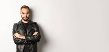 Deri ceket giyen yakışıklı bir adam, kameraya özgüvenli bakıyor, beyaz arka planda duruyor..