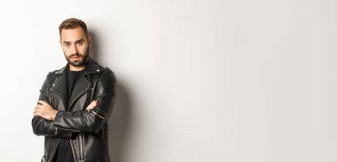 Çekici, sakallı, deri ceketli, gülümseyen, elleri göğsünde çaprazlanmış, beyaz arka planı olan maço bir adam..