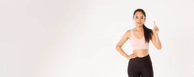 Spor, refah ve aktif yaşam tarzı konsepti. Gülümseyen Asyalı fitness eğitmeni, spor giyim dersindeki kız, birinci kuralı gösteriyor, parmağını kaldır ve gülümse, beyaz arka planda dur..