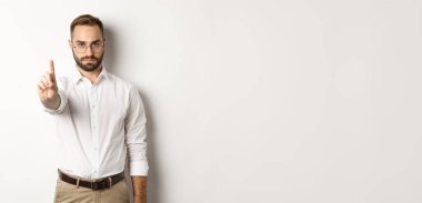 Gözlüklü ciddi bir adam dur işareti gösteriyor, yasaklamak ve yasaklamak için parmağını sallıyor, beyaz arka planda duruyor..