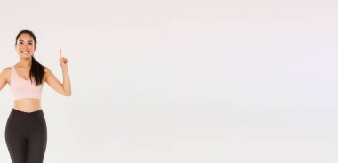 Spor kıyafetleri içinde mükemmel vücut yapısı olan yakışıklı Asyalı bir kız, sporu seven bir bayan sporcu, parmaklarını kaldırıp idman ekipmanlarıyla reklamlara bakıyor, beyaz arka plan..