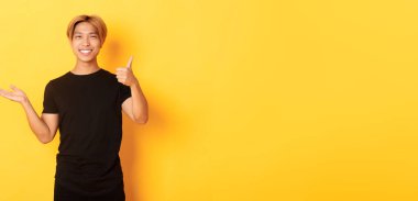 Memnun ve mutlu çekici Koreli adam gülümsüyor, sevinçle onaylıyor, elinde bir şey tutuyor, sarı arka planda duruyor..