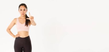 Spor, refah ve aktif yaşam tarzı konsepti. Ciddi derecede hoşnutsuz Asyalı kadın fitness eğitmeni, sporcu kadın onaylamayarak, uyararak ya da karşı çıkarak parmağını sallıyor, beyaz arka planda duruyor..