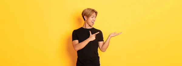 Portrét Pohledného Usměvavého Asiata Držícího Něco Ruce Ukazujícího Prstem Stojící — Stock fotografie