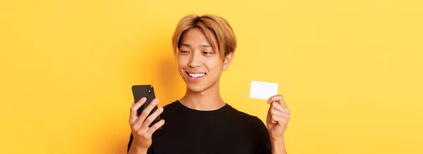 携帯電話と笑顔を見てクレジットカードを見せ黄色の背景の上に立って — ストック写真