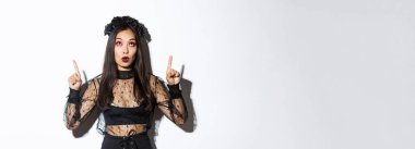 Cadı kılığındaki çekici Asyalı kadın Cadılar Bayramı bayrağını ve parti tanıtımını işaret ediyor. Siyah gotik elbiseli güzel bir kadın. Şaşırmış görünüyor..