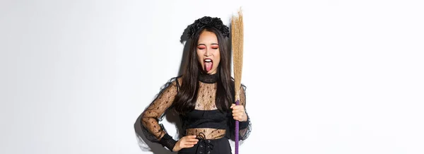 无忧无虑的亚洲女人 享受派对之间的光环 穿着巫婆服装 拿着扫帚 露出舌头 站在白色的背景上 — 图库照片