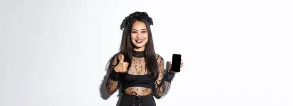 照片中 身穿哈洛洛人服装的快乐而满意的亚洲女人露出大拇指和展示手机屏幕 高兴地微笑着 站在白色的背景上 — 图库照片